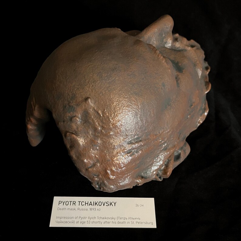 Puede incluir: Una m&aacute;scara mortuoria de color bronce de Piotr Chaikovski, creada en Rusia en 1893. La m&aacute;scara mide aproximadamente 26 cm de largo. La imagen incluye una etiqueta con el nombre del compositor y detalles sobre la creaci&oacute;n de la m&aacute;scara.