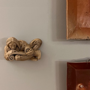 May include: A beige-colored relief sculpture of a person reclining and reading a scroll. The figure is detailed with flowing hair and clothing. The sculpture is mounted on a gray wall, with two framed artworks in the background.