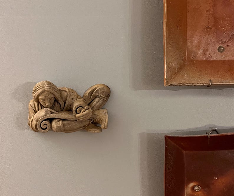 May include: A beige-colored relief sculpture of a person reclining and reading a scroll. The figure is detailed with flowing hair and clothing. The sculpture is mounted on a gray wall, with two framed artworks in the background.