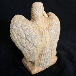 May include: A stone sculpture of a bird with outstretched wings. The sculpture is a light beige color with detailed feather carvings. The bird is perched on a rectangular base, set against a black background. The sculpture is viewed from the back.