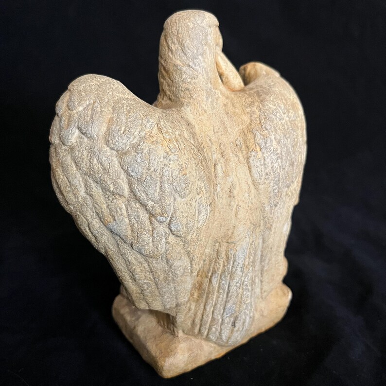 May include: A stone sculpture of a bird with outstretched wings. The sculpture is a light beige color with detailed feather carvings. The bird is perched on a rectangular base, set against a black background. The sculpture is viewed from the back.