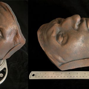 May include: A bronze-colored sculpture of a human face, with closed eyes, resting on a digital scale displaying 474. A ruler shows measurements in inches. The sculpture is a bust, with the head and upper neck visible.