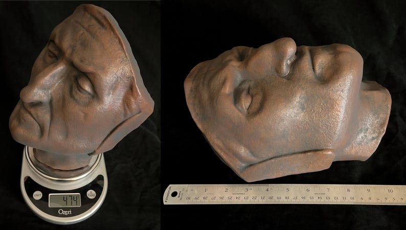 May include: A bronze-colored sculpture of a human face, with closed eyes, resting on a digital scale displaying 474. A ruler shows measurements in inches. The sculpture is a bust, with the head and upper neck visible.