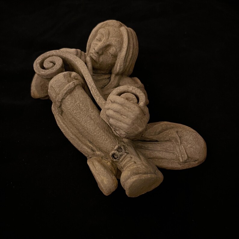 May include: A beige-colored sculpture of a person seated with a curved object. The figure is wearing a hooded garment and holding a round object. The sculpture has a textured surface and is set against a black background.