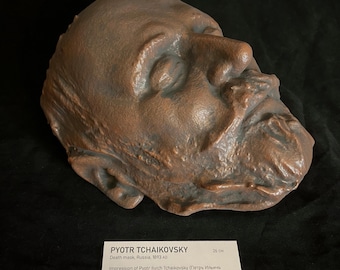 May include: A bronze-colored death mask of Pyotr Tchaikovsky, created in Russia in 1893. The mask is a detailed impression of the composer's face, with visible facial features. A label below indicates the mask is 26 cm in length.