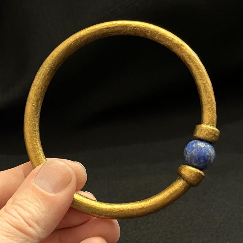 May include: A gold-coloured bangle bracelet with a textured surface. The bracelet features a round, dark blue stone set into the design. The bracelet is held against a black background.