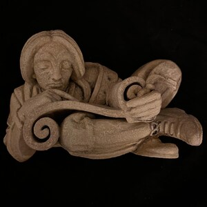 May include: A beige-colored sculpture of a person seated and reading a scroll. The figure is wearing a long-sleeved garment and has their eyes closed. The scroll is held in both hands and has a decorative curled end.