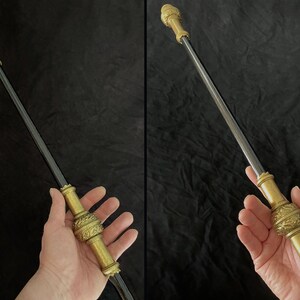 May include: An ornate walking stick with a dark shaft and gold-coloured metal detailing. The top has a detailed sculpture, with intricate designs on the handle and midsection. The walking stick is held in a person's hand.