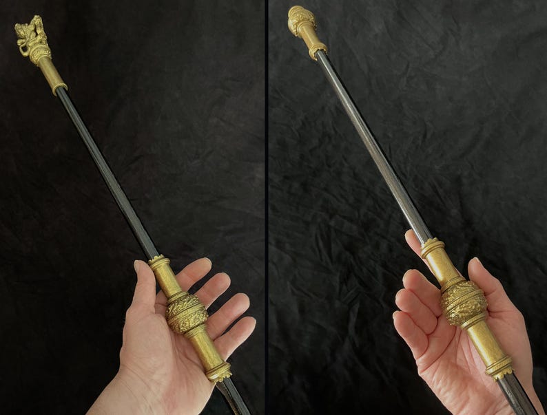 May include: An ornate walking stick with a dark shaft and gold-coloured metal detailing. The top has a detailed sculpture, with intricate designs on the handle and midsection. The walking stick is held in a person's hand.