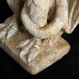 May include: A detailed view of a gargoyle statue, showcasing its textured, weathered appearance. The sculpture features a clawed foot resting on a square base, with a wing and arm visible. The color palette is primarily beige and brown, with a focus on the intricate details.