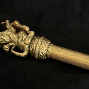 May include: A gold-coloured staff featuring a detailed lion figure at the top. The staff has a cylindrical design with a black handle. The lion is in a reclining position, with intricate details.