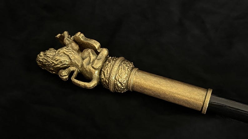 May include: A gold-coloured staff featuring a detailed lion figure at the top. The staff has a cylindrical design with a black handle. The lion is in a reclining position, with intricate details.