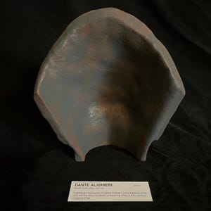 May include: A weathered, gray-blue sculpted effigy, possibly a death mask, with a curved, open form. The surface shows hints of brown and rust-like coloration. The image includes a label with the text "DANTE ALIGHIERI" and measurements of 26 cm.
