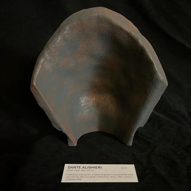 May include: A weathered, gray-blue sculpted effigy, possibly a death mask, with a curved, open form. The surface shows hints of brown and rust-like coloration. The image includes a label with the text "DANTE ALIGHIERI" and measurements of 26 cm.