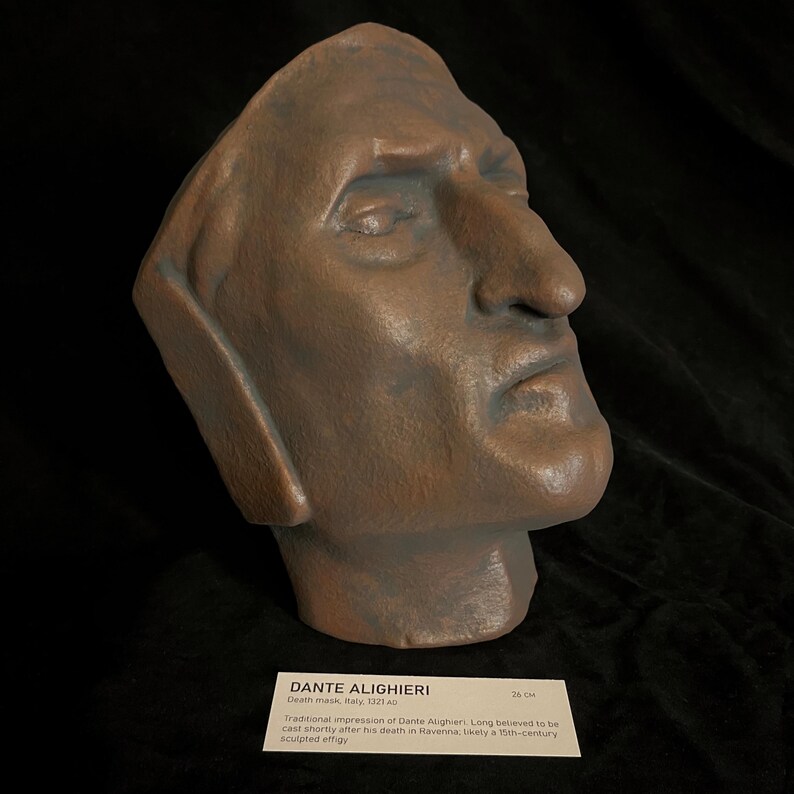 May include: A bronze-colored sculpted death mask of Dante Alighieri. The mask is a side profile with a prominent nose and detailed facial features. A label reads "DANTE ALIGHIERI Death mask, Italy, 1321 AD" and "26 CM".