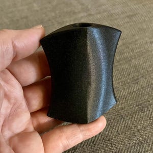 Puede incluir: Una talla de piedra negra, lisa y pulida, con forma de escudo y un peque&ntilde;o agujero en la parte superior.