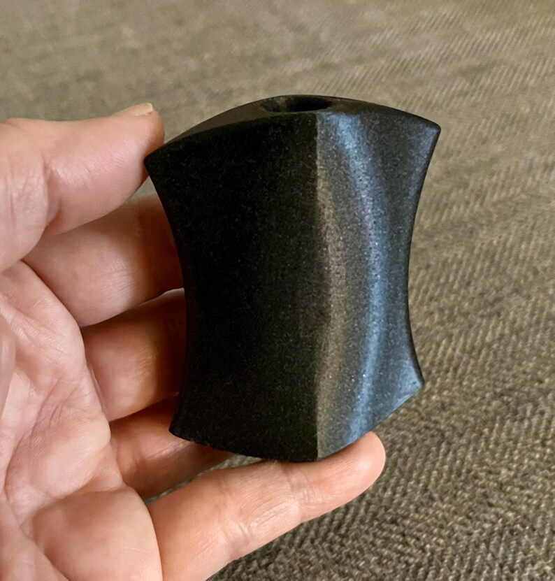 Puede incluir: Una talla de piedra negra, lisa y pulida, con forma de escudo y un peque&ntilde;o agujero en la parte superior.
