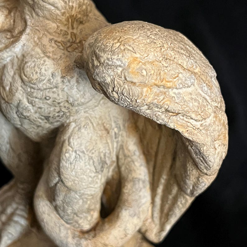 May include: A close-up of a weathered, off-white sculpture of a winged creature. The sculpture has a textured surface with visible details in the wings and body. The background is dark, highlighting the sculpture's form.