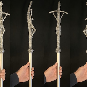 May include: A silver-colored religious staff featuring a detailed depiction of the crucifixion. The staff has a textured, ornate design with a figure of Jesus on the cross. The staff is held by a person.