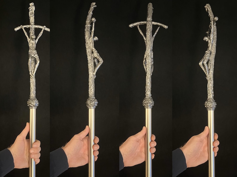 May include: A silver-colored religious staff featuring a detailed depiction of the crucifixion. The staff has a textured, ornate design with a figure of Jesus on the cross. The staff is held by a person.
