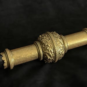 May include: Close-up of a decorative staff or sceptre. The central section is a detailed, ornate gold colour with a textured, floral design. The ends are also gold, with a simpler, banded design. The shaft is a dark, metallic colour.