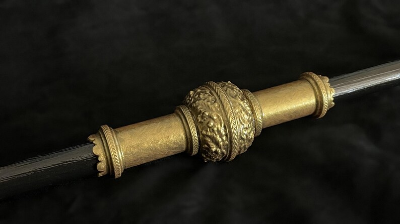 May include: Close-up of a decorative staff or sceptre. The central section is a detailed, ornate gold colour with a textured, floral design. The ends are also gold, with a simpler, banded design. The shaft is a dark, metallic colour.