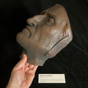 May include: A brown sculpted death mask of Dante Alighieri, with a detailed profile. The mask is held by a hand. A small white card with text is visible below the mask, including the name "DANTE ALIGHIERI".