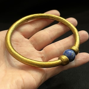 May include: A gold-coloured bangle bracelet with a hammered texture. The bracelet features a large, round, blue gemstone bead. The bracelet is held in a hand against a dark background. Jewellery.