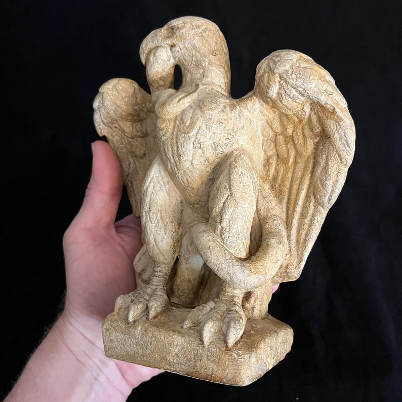 May include: A beige-colored statue of a griffin, a mythical creature with the body of a lion and the head and wings of an eagle. The statue has detailed feather and wing textures, with a curled tail and is mounted on a rectangular base.