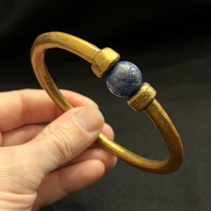 May include: A gold-coloured metal bracelet featuring a central blue sphere. The bracelet has a curved, open design with a thick band. The blue sphere is held in place by two rectangular metal pieces. The bracelet is held in a hand.