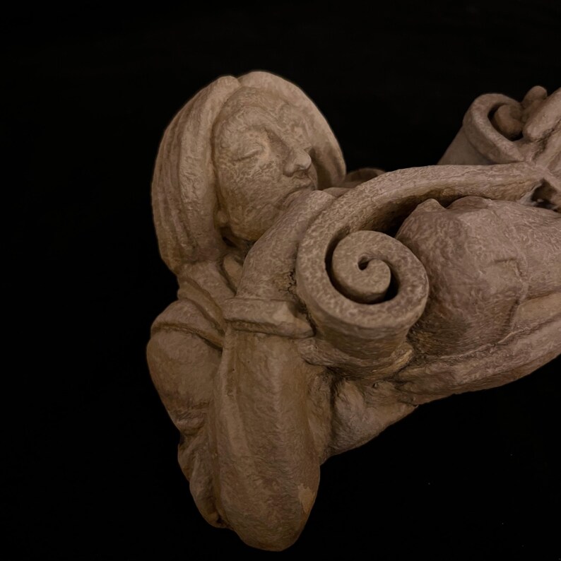 May include: A detailed, close-up view of a stone sculpture featuring a woman's face with closed eyes, intertwined with a swirling, scroll-like design. The sculpture is a muted brown color, set against a black background.