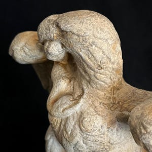 May include: A detailed sculpture of a figure with a textured, off-white surface. The sculpture has a rough, aged appearance with visible details and shadows. The figure's form is partially visible against a dark background, highlighting its shape and texture.