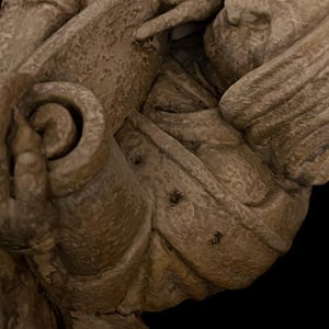 May include: Close-up of a weathered, beige sculpture fragment. The image highlights a detailed, curved scroll design and textured surfaces, possibly part of a larger architectural or decorative piece. The sculpture has a rough, aged appearance.