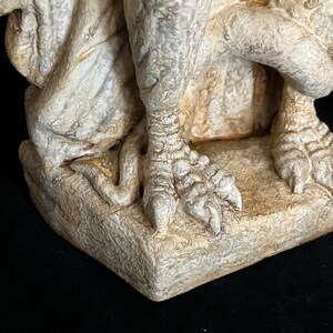 May include: Close-up of a weathered, off-white gargoyle statue. The image focuses on the detailed clawed feet and textured base, showcasing the sculpture's aged appearance and intricate design. The statue has a rough, stone-like texture.