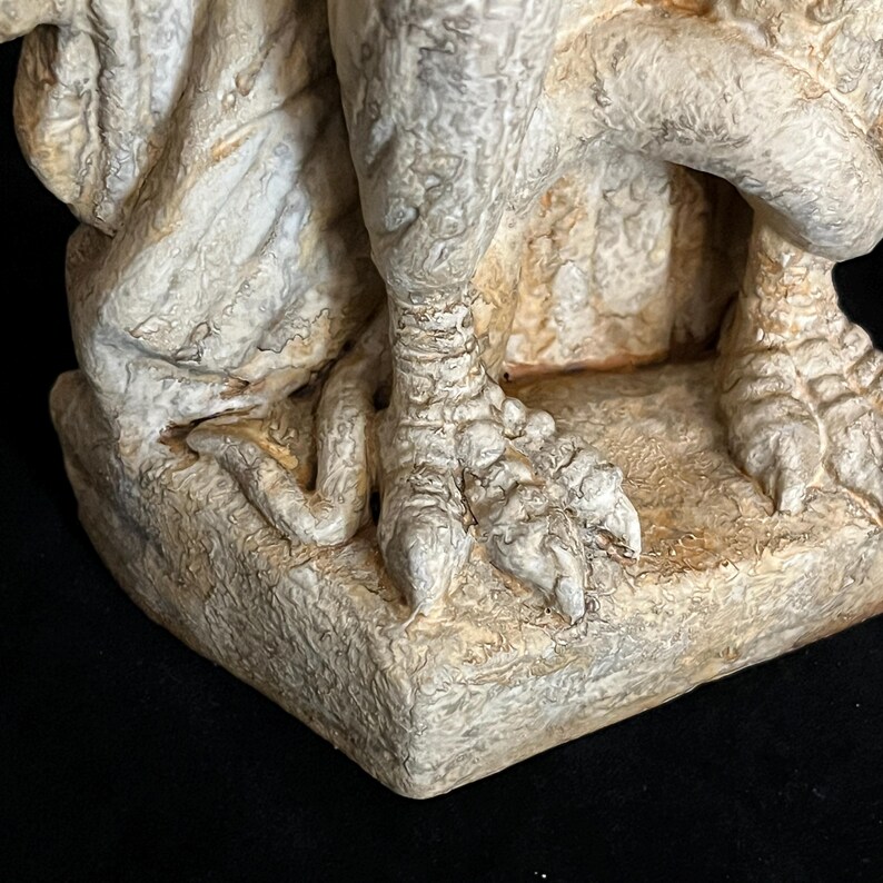 May include: Close-up of a weathered, off-white gargoyle statue. The image focuses on the detailed clawed feet and textured base, showcasing the sculpture's aged appearance and intricate design. The statue has a rough, stone-like texture.
