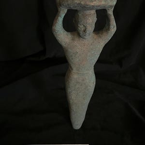 May include: A green bronze Sumerian foundation peg, a ceremonial object buried beneath temples to sanctify sacred spaces and invoke divine protection and royal authority. It depicts a figure carrying a large object on their head. The peg is 22 cm tall.