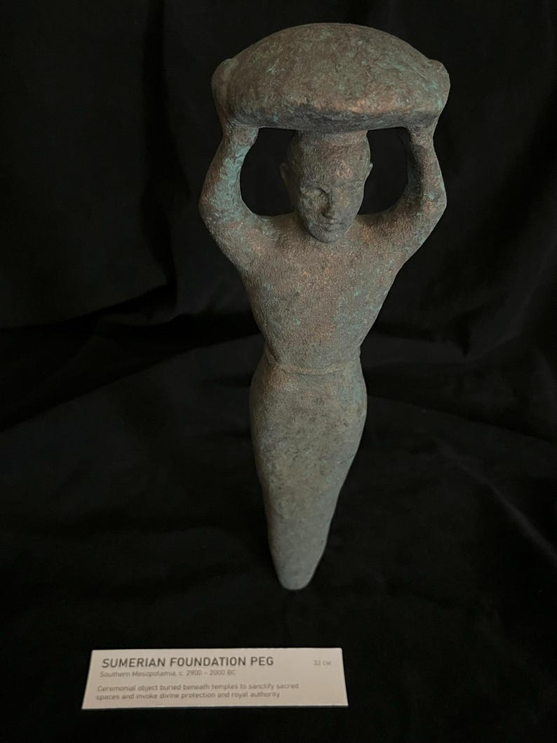 May include: A green bronze Sumerian foundation peg, a ceremonial object buried beneath temples to sanctify sacred spaces and invoke divine protection and royal authority. It depicts a figure carrying a large object on their head. The peg is 22 cm tall.