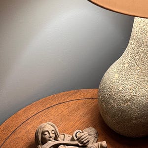 May include: A carved stone sculpture of a person playing a stringed instrument, lying down. The sculpture is on a wooden table, with a textured lamp base and lampshade in the background.