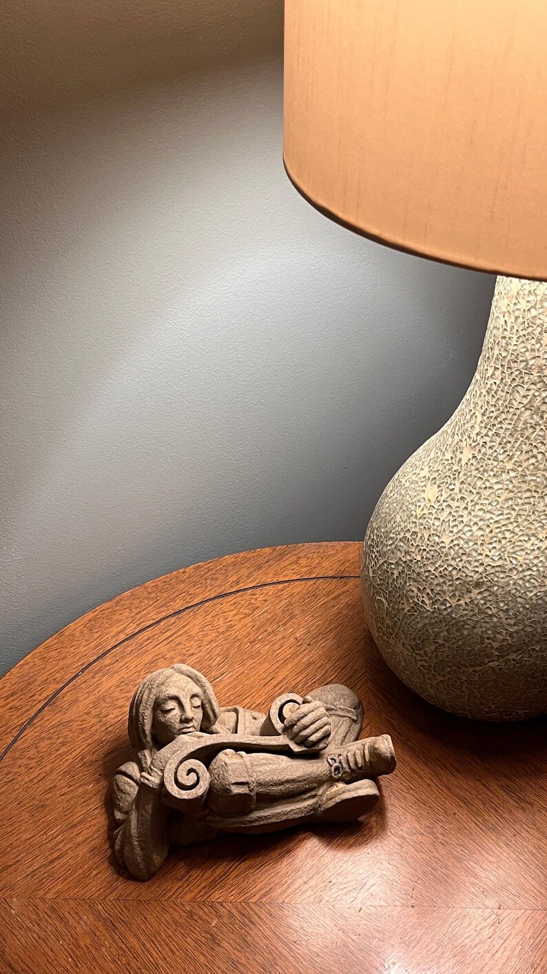 May include: A carved stone sculpture of a person playing a stringed instrument, lying down. The sculpture is on a wooden table, with a textured lamp base and lampshade in the background.
