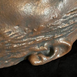 Puede incluir: Primer plano de una escultura texturizada de color bronce con una capa base gris oscuro. La superficie tiene un aspecto rugoso y org&aacute;nico con sutiles variaciones de color y textura. La escultura tiene una forma redondeada con una peque&ntilde;a hendidura curva.