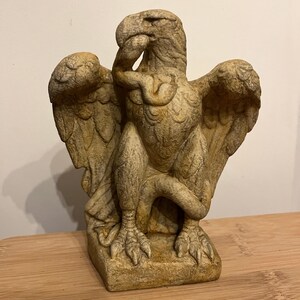 May include: A detailed, weathered statue of an eagle with outstretched wings. The sculpture is in a seated position, with its head tilted and beak touching its wing. The eagle is a light tan color, with textured details on the feathers and body.