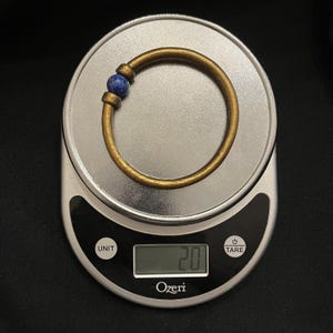 May include: A brass bangle with a blue gemstone bead. The bracelet is on a digital scale, which reads 20. The bracelet is circular and has a simple design.