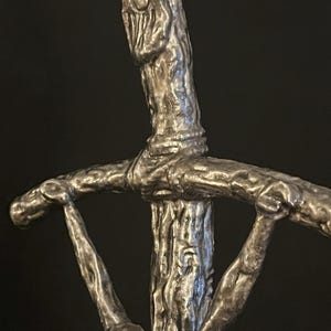 May include: A close-up of a silver-colored sword hilt with a textured, tree-like design. The hilt features a crossguard with stylized arms and a detailed, organic appearance. The sword's design has a rustic, antique aesthetic.