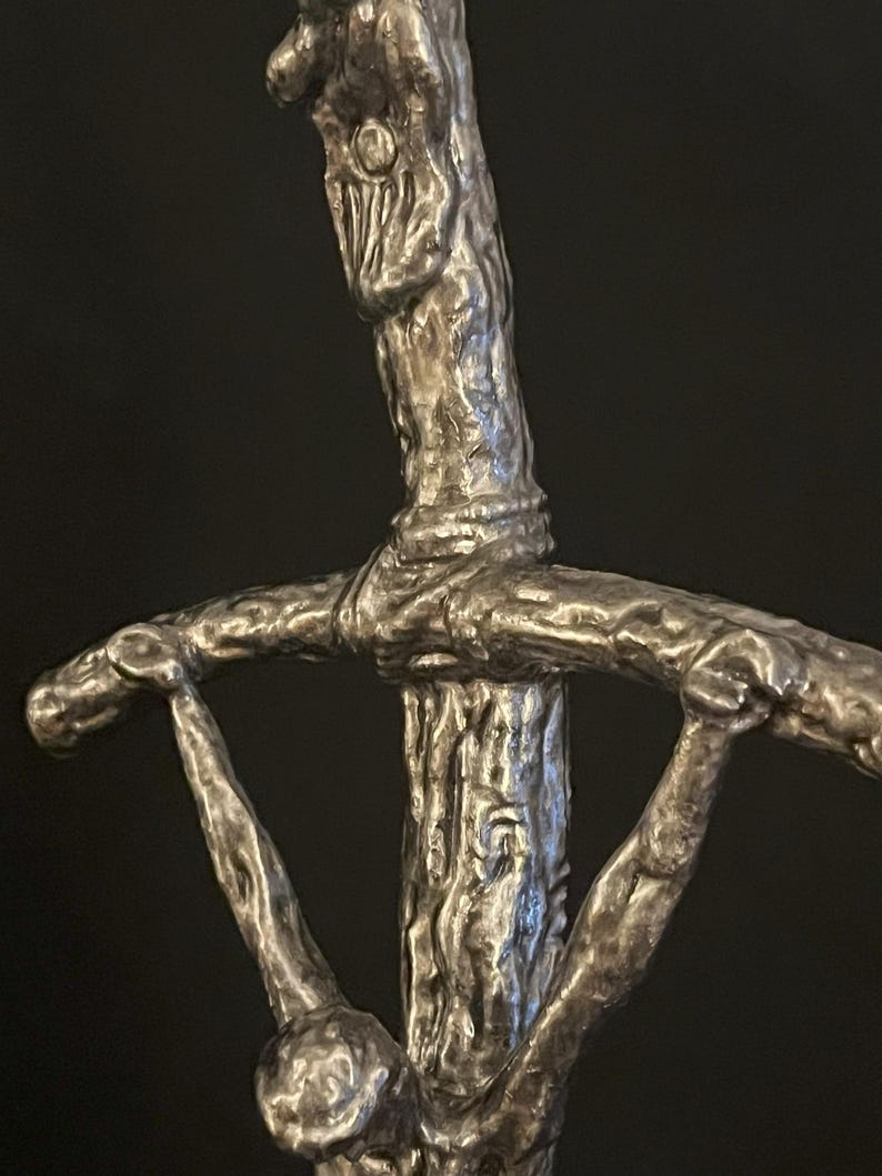 May include: A close-up of a silver-colored sword hilt with a textured, tree-like design. The hilt features a crossguard with stylized arms and a detailed, organic appearance. The sword's design has a rustic, antique aesthetic.