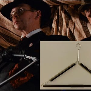 May include: A metal and black spring-loaded clothes hanger. The hanger has a silver hook and black bars. The image also shows a scene from a movie with two men in suits and hats.