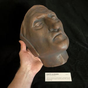 May include: A bronze-colored death mask of Dante Alighieri, with closed eyes and a serene expression. The mask is held in a hand, and a label below reads "Dante Alighieri Death mask, Italy, 1321 AD". The mask measures 26 cm.