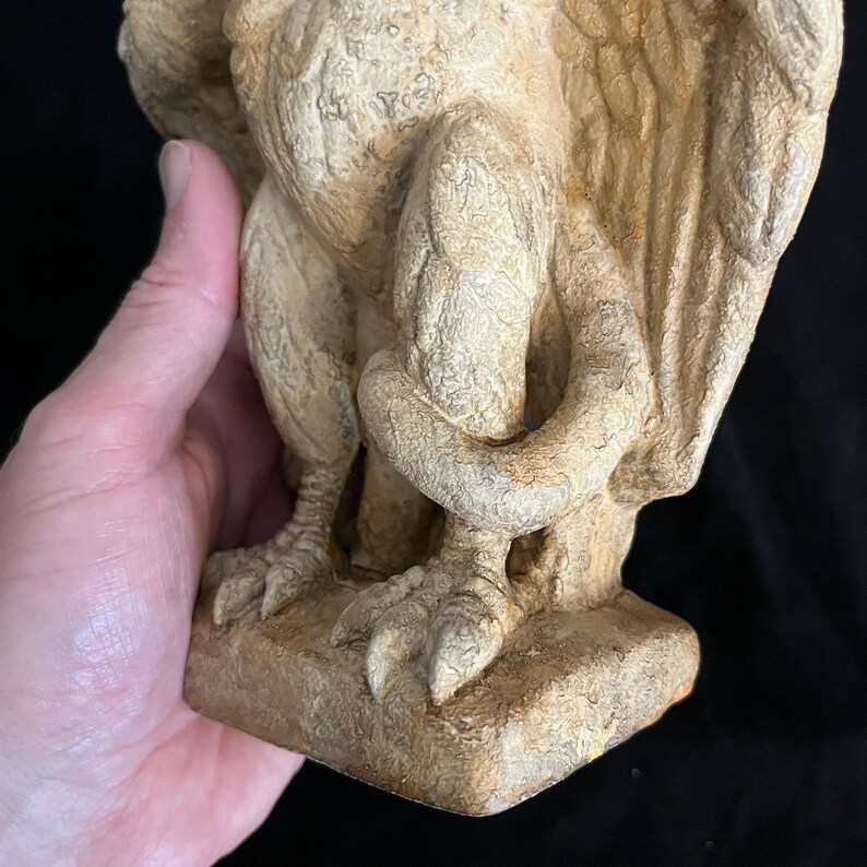 May include: A detailed view of a beige gargoyle statue. The statue features textured wings, clawed feet, and a curled tail. The gargoyle is resting on a square base, with visible details in the stone.