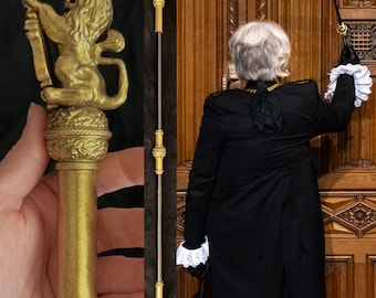 May include: A gold-colored ceremonial staff with a lion figure at the top. The staff is held by a person wearing a black coat with white lace cuffs, standing in front of a wooden door.