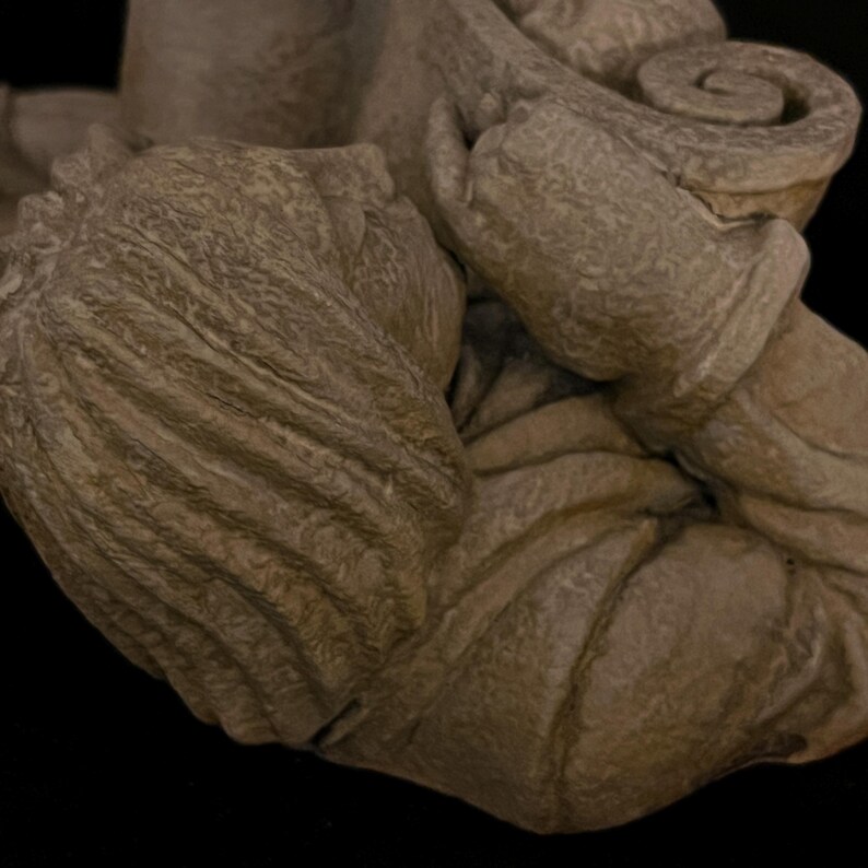 May include: Close-up of a weathered, stone-like sculpture with a textured surface. The sculpture features detailed folds and curves, with a focus on the intricate design of the piece. The color is a muted, earthy tone.