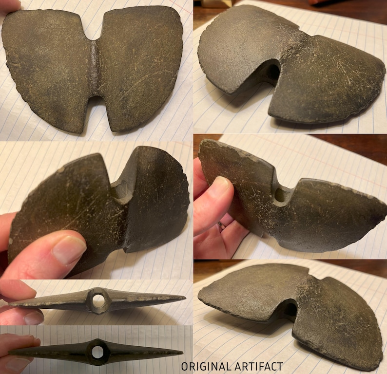 May include: A dark gray stone artifact with a unique shape, featuring a hole in the center and a split on one side. The artifact is likely an ancient tool or weapon.